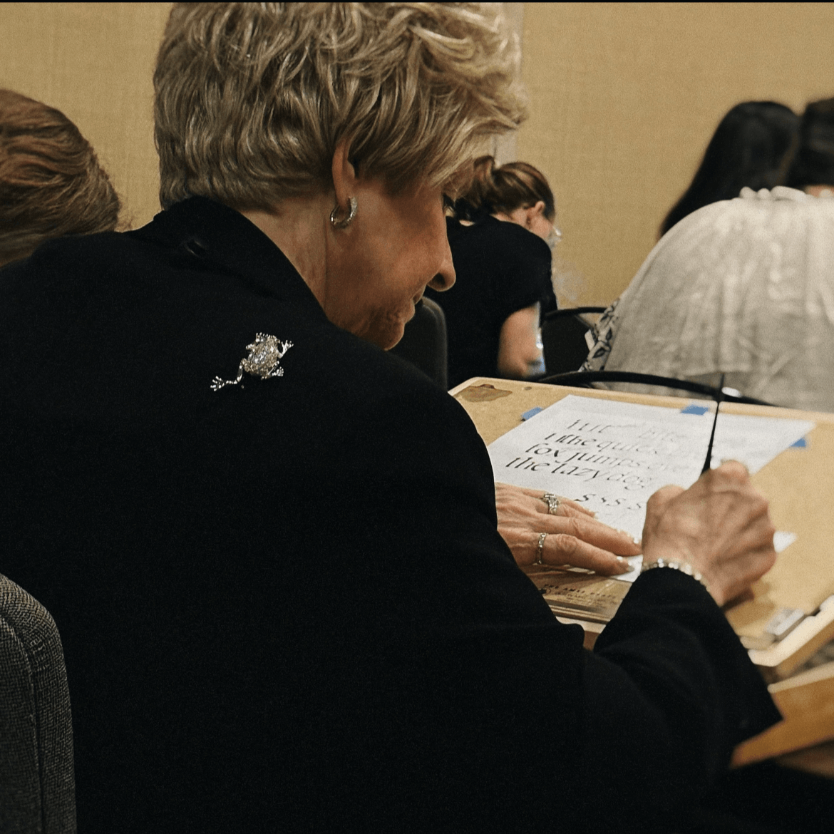 A focused elderly woman with short blonde hair, dressed in a black outfit, writing on paper during a group activity, possibly a workshop or class. She is adorned with a diamond-encrusted frog-shaped brooch, alongside a ring and bracelet, adding a touch of elegance and personality to her attire. The setting is a typical indoor environment with participants gathered around a table, indicating a collective learning or creative activity. The frog brooch, often symbolizing luck and vitality, serves as a distinctive accessory reflecting the wearer's refined taste.
