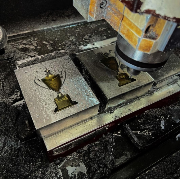 CNC milling machine engraving a trophy-shaped metal mold, showing completed positive mold and cutting in progress on the negative mold, used for custom enamel pin and badge production