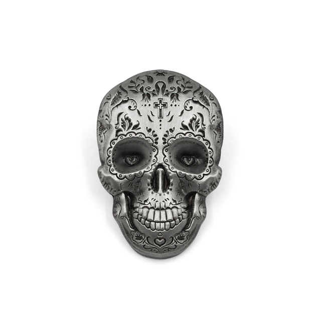 Close-up of an antiqued silver die cut metal pin featuring a detailed sugar skull design. The pin shows intricate floral engravings, heart-shaped eyes, and a cross on the forehead with a 3D relief effect