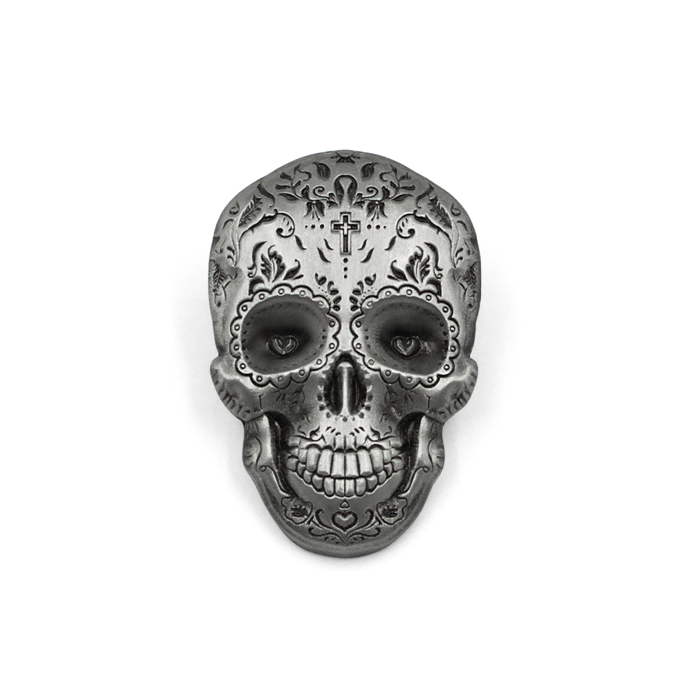 Close-up of an antiqued silver die cut metal pin featuring a detailed sugar skull design. The pin shows intricate floral engravings, heart-shaped eyes, and a cross on the forehead with a 3D relief effect
