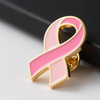 A close-up of a pink ribbon lapel pin on a blurred dark background, emphasizing the hard enamel filling and metal edging. The pin is a symbol of Breast Cancer Awareness, commonly used in dedicated awareness events, with a clear focus on its elegant design and meaningful caus