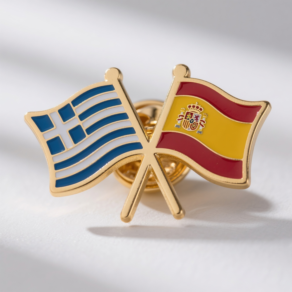 Two enamel lapel pins in a crossed display, featuring the flags of Greece and Spain. The Greek flag shows blue and white stripes with a blue cross, while the Spanish flag features red and yellow stripes with the Spanish coat of arms. Both pins have metal edging and enamel filling, often used in cultural exchanges, international events, or as souvenirs between the two countries.