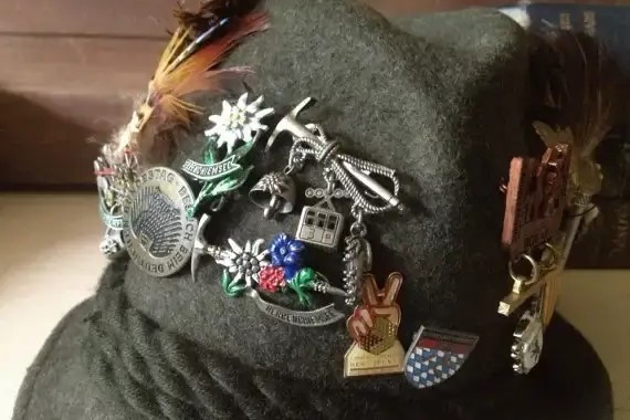 Close-up of a felt hat adorned with multiple metal souvenir pins and charms, including flowers, tools, and emblems.