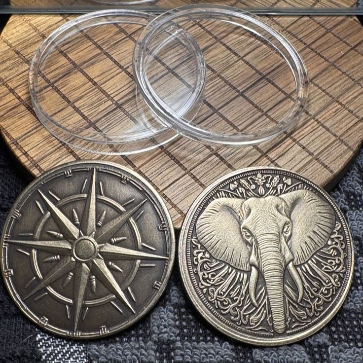 Two antique-style metal collectible coins featuring a detailed compass rose and ornate elephant design, displayed with clear protective cases for travel souvenirs and cultural gift collections