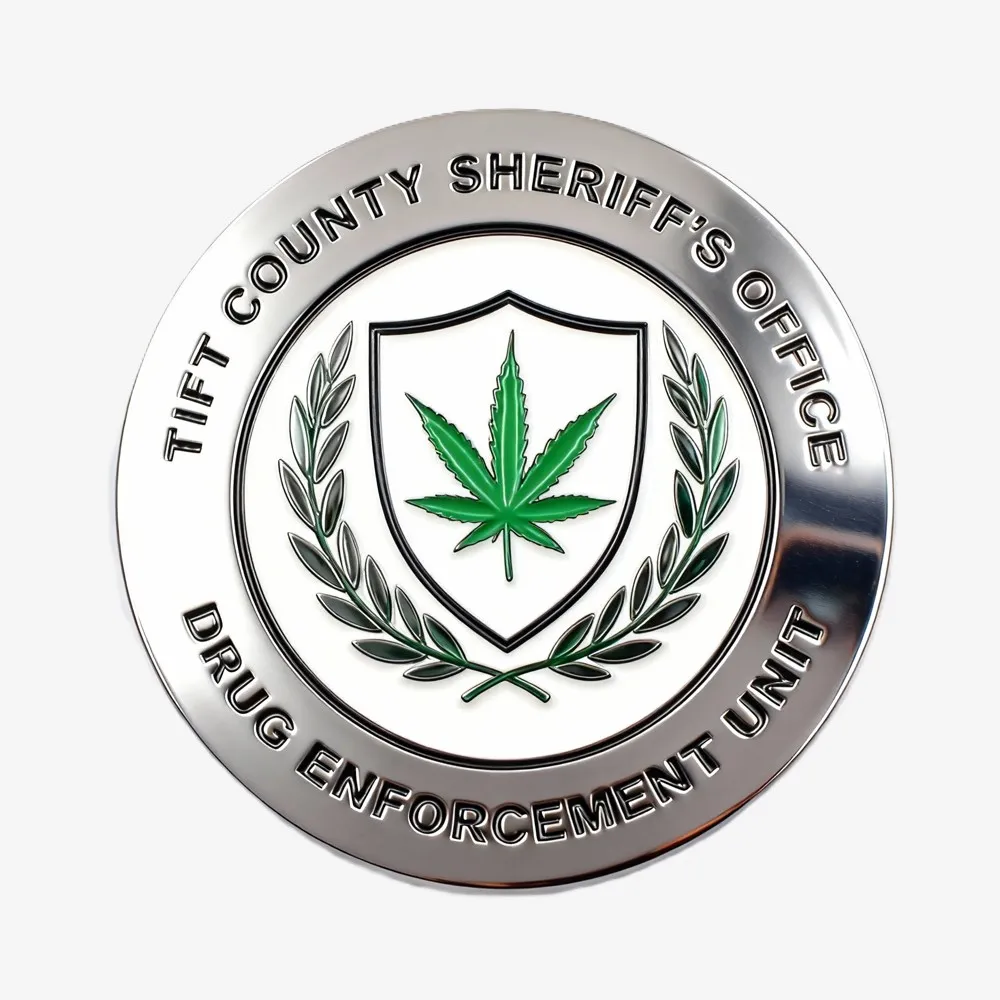 Silver round sheriff’s office drug enforcement challenge coin with a shield, green leaf, and laurel branch design in the center.